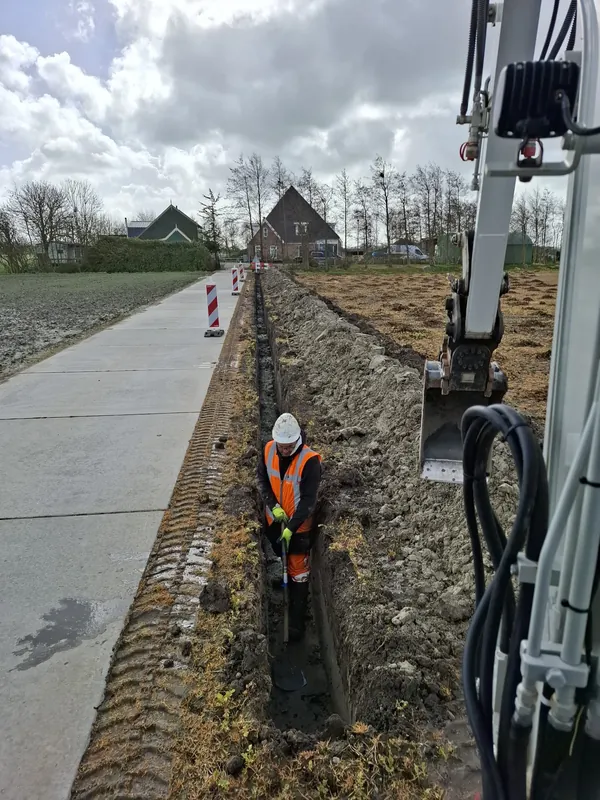 Grondwerker graaft met een schop in een sleuf langs een weg, naast een minigraver